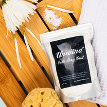 Load image into Gallery viewer, Package of 'Unwind Bath Fizzy Dust' with a yellow bath sponge on a wooden surface.