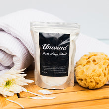 Load image into Gallery viewer, Bath fizzy dust package labeled 'Unwind' with a sponge and flowers on a wooden surface.