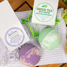 Load image into Gallery viewer, Two bath bombs, one green and one purple, with their packaging on a white towel.