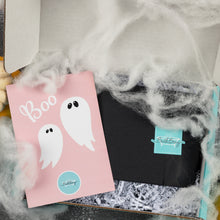 Load image into Gallery viewer, A Halloween-themed gift box from Bath Bevy featuring a pink card with cute white cartoon ghosts and the word 'Boo', next to a black envelope with a turquoise logo, surrounded by fake white spiderwebs and shredded paper.