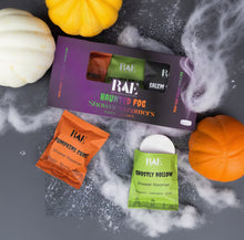 Load image into Gallery viewer, RAE brand haunted fog shower steamers gift set with three sealed packets (Pumpkin Curse, Cherry Hollow, Salem) arranged around an open purple box on a dark textured surface with white faux fog and pumpkin props.