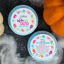 Load image into Gallery viewer, Two round blue 'Wake the Dead' Peppermint shower steamers are arranged on a dark textured surface with white faux cobwebs and part of an orange pumpkin visible. The top tub lid shows a white label with colorful skull illustrations and the product name, while the bottom tub shows the full ingredients list.