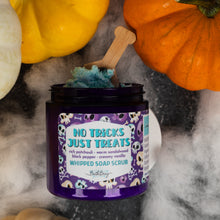 Load image into Gallery viewer, A jar of whipped soap scrub labeled 'No Tricks Just Treats' sits on a dark surface with Halloween-themed props including pumpkins and white fog. The jar is open, showing blue whipped scrub, with a small wooden spoon resting inside.
