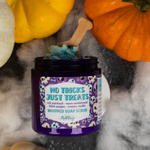 Load image into Gallery viewer, A jar of whipped soap scrub labeled 'No Tricks Dust Treats' sits on a dark surface with Halloween-themed props including pumpkins and white fog. The jar is open, showing blue whipped scrub, with a small wooden spoon resting inside.