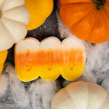 Load image into Gallery viewer, A wavy candy corn-shaped bath bomb with a yellow, orange, and white horizontal stripe, arranged on a dark surface with white faux cobwebs and decorative pumpkins.