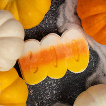 Load image into Gallery viewer, A Halloween-themed bath bomb shot featuring a candy corn-shaped or candle in yellow, orange, and white, surrounded by decorative miniature pumpkins and white cobweb decorations on a dark textured background.
