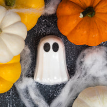 Load image into Gallery viewer, A white ghost-shaped bath bomb with black oval eyes, styled on a dark surface with orange and white pumpkins and white cobweb-like decoration.