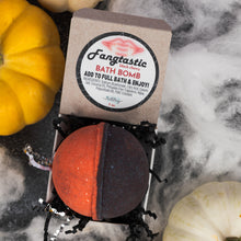 Load image into Gallery viewer, A round bath bomb in an open box with the label 'Fangtastic Black Cherry Bath Bomb' visible, styled with small pumpkins and white cobweb decor on a dark surface.