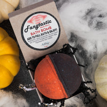 Load image into Gallery viewer, A Fangtastic Black Cherry bath bomb in its kraft paper packaging, styled with autumn props including a yellow pumpkin, a white pumpkin, and cobwebs on a dark surface.