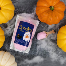 Load image into Gallery viewer, A package of pink 'Spooky Fizzy Dust' sits on a dark gray surface surrounded by decorative pumpkins and white faux cobwebbing. A small wooden scoop filled with the pink bath salts is placed to the right of the package.