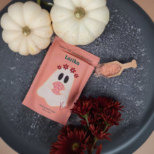 Load image into Gallery viewer, A pink packet of Latika Boo Bath Soak featuring a ghost illustration, styled on a dark gray textured surface with white pumpkins, red flowers, and a wooden scoop of bath soak.