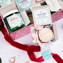 Load image into Gallery viewer, A holiday-themed 12 Days of Bathmas bath gift set featuring a pink box with multiple bath products, including a green jar of 'Holiday Garland' whipped soap scrub and a bath bomb in an open box. The scene is decorated with red velvet ribbon, red berries, and candy canes on a white background.