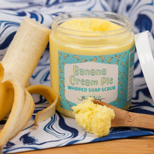 Load image into Gallery viewer, Jar of Banana Cream Pie Whipped Soap Scrub with a spoonful of scrub and banana peels on a blue and white patterned cloth.