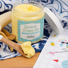 Load image into Gallery viewer, Jar of Banana Cream Pie whipped soap scrub with a wooden spoon and banana on a blue and white patterned cloth.