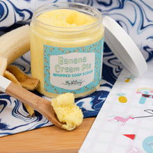 Load image into Gallery viewer, Jar of Banana Cream Pie whipped soap scrub with a wooden spoon and banana on a blue and white patterned cloth.