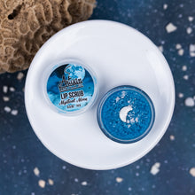 Load image into Gallery viewer, small round jar of lip scrub from Artisan, labeled 'Mystical Moon' in blue, arranged on a white ceramic plate against a dark blue speckled background with a coral prop.
