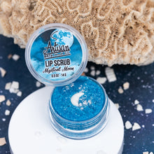 Load image into Gallery viewer, An open jar of blue lip scrub with a matching lid leaning above it, styled on a white round plate with a coral-like prop and white shell pieces on a dark blue background. The lid label reads Artisan Bath Co.