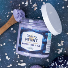 Load image into Gallery viewer, A jar of Bath Bevy 'Starry Night' whipped soap scrub with a white lid off, showing the lavender scrub inside. A wooden spoon with scrub is on the left, and decorative stones are scattered on a dark blue speckled background.