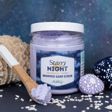 Load image into Gallery viewer, A jar of whipped soap scrub labeled 'Starry Night' by Bath Bevy, filled with light purple whipped soap scrub, shown on a dark surface with a wooden-handled spatula holding some scrub, a sponge, and decorative stones against a blue speckled background.