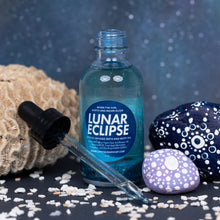Load image into Gallery viewer, A bottle of Lunar Eclipse Crystal Infused Bath and Body Oil with a glass dropper, styled with natural coral, white shells, and patterned stones against a dark blue speckled background.