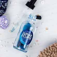 Load image into Gallery viewer, A clear glass dropper bottle labeled 'LUNAR ECLIPSE' with blue liquid inside, arranged on a white surface with scattered shell fragments and decorative stones. The dropper and cap are placed nearby.