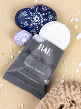 Load image into Gallery viewer, A packaged shower steamer from RAE The Artisanal Shop, labeled 'Moon Magic' with lavender and citrus notes, shown with a white round steamer cap on a beige surface with decorative stones and seashells.