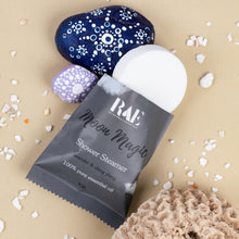 Load image into Gallery viewer, A packaged shower steamer from RAE Home &amp; Body Essentials labeled 'Moon Magic' lies on a beige surface with decorative stones and small shell pieces around it. The product itself is partially visible as a white round pad or packet insert.