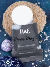 Load image into Gallery viewer, A 1 oz packet of RAE Moon Magic Shower Steamer essential oil product, shown with a white round steamer cap resting on top, styled with spa props on a dark blue textured background.