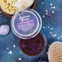 Load image into Gallery viewer, A top-down styled product shot of a round tub of Loofah Soap labeled 'Galactic Grape'. The clear lid shows a dark purple and black patterned label, and the soap inside is a deep purple.