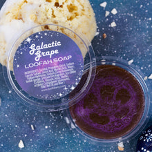 Load image into Gallery viewer, Galactic Grape Loofah Soap, showing the deep purple loofah inside, with a decorative label on top.  on a textured blue surface.