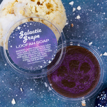 Load image into Gallery viewer, A clear plastic tub of Galactic Grape Loofah Soap by Bath Bevy, showing the deep purple soap with loofah inside, with a decorative label on top. background on a textured blue surface.